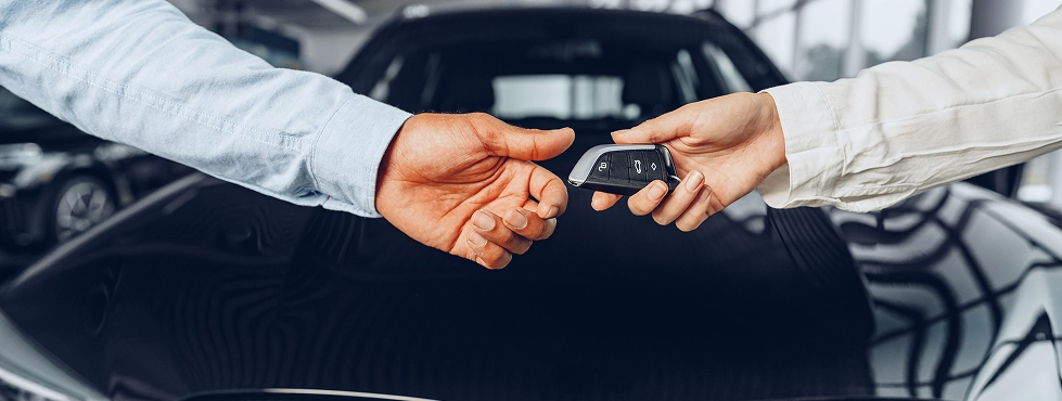 Twee mensen wisselen autosleutels uit, met op de achtergrond een zwarte luxeauto, wat symbool staat voor een transactie of de aankoop van een auto in een lichte showroom.