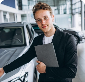 Een glimlachende verkoper in een zwart pak houdt een map vast en wijst naar een zilverkleurige auto in een lichte, moderne showroom, waarbij hij een professionele en gastvrije indruk maakt.